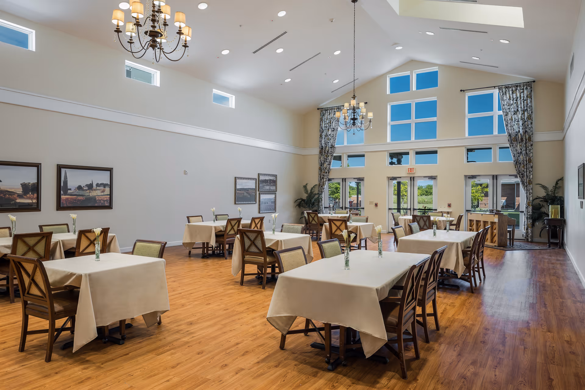 Spacious dining room with multiple tables covered in beige tablecloths, wooden chairs, chandeliers, and large windows letting in natural light.