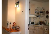 Interior view showing a small kitchen area with light wood cabinets, a microwave, and various kitchen items. To the left, there is a small ledge with decorative figurines and a wall-mounted light fixture above it.