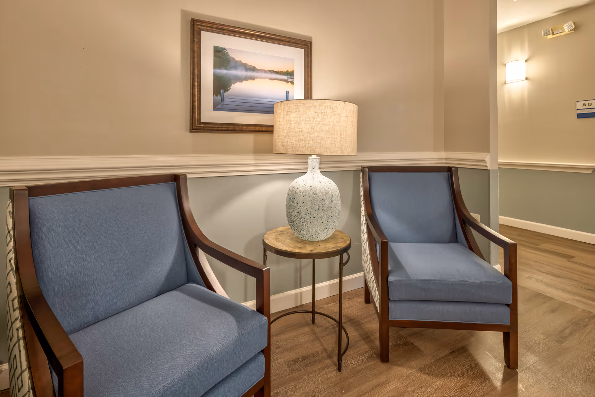 Two blue cushioned wooden armchairs with a small round wooden side table between them holding a ceramic table lamp with a beige shade. A framed picture of a dock on a lake at sunrise hangs on the beige wall behind the chairs. The floor is wood, and there is a hallway visible to the right with a wall sconce light and a room number sign.