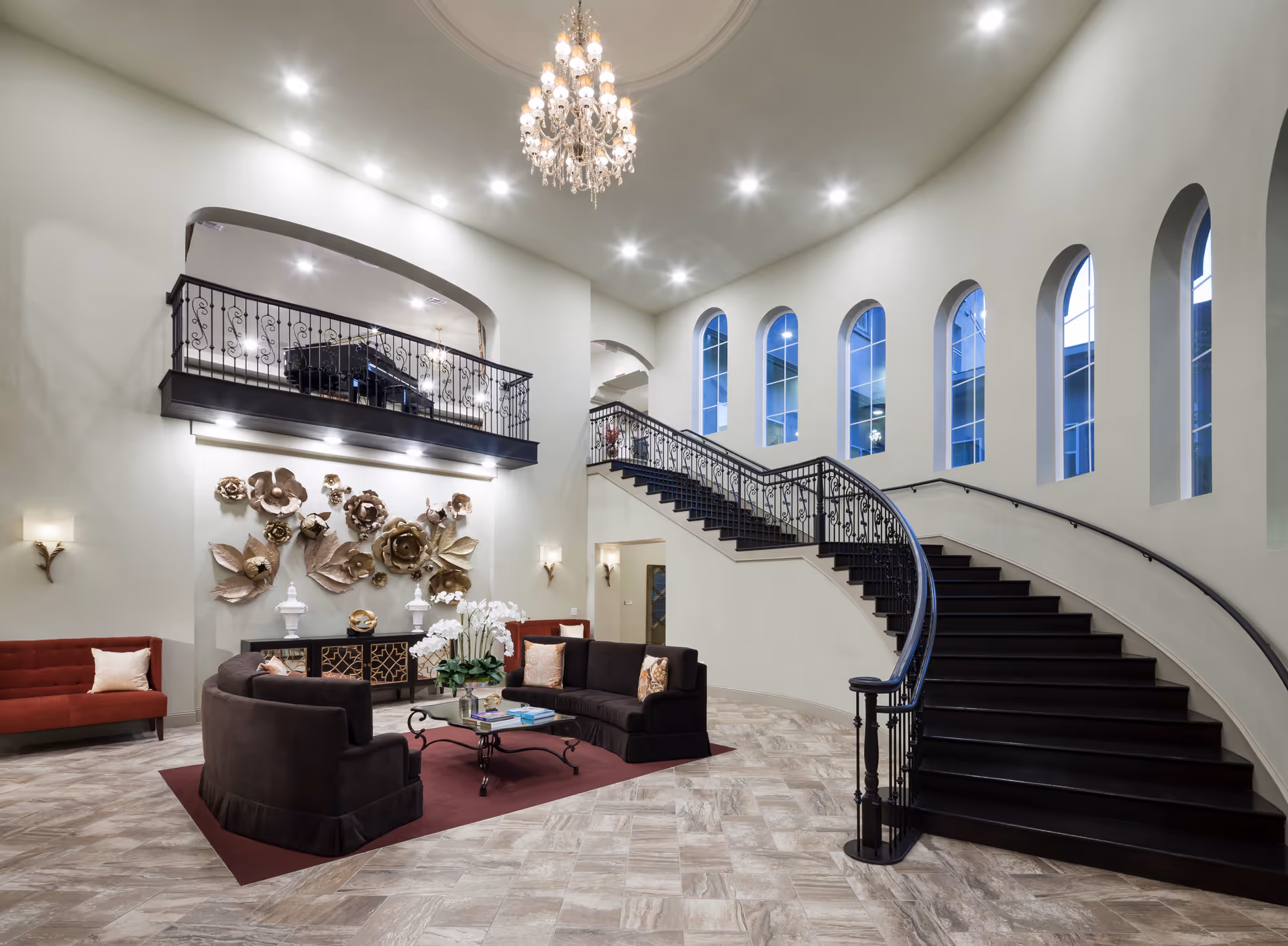 Spacious and elegant interior of a senior living facility featuring a curved black staircase with ornate railing, a chandelier hanging from the ceiling, tall arched windows letting in natural light, a seating area with dark sofas and a red rug, decorative wall art with large metallic flowers, and a red bench with a pillow.