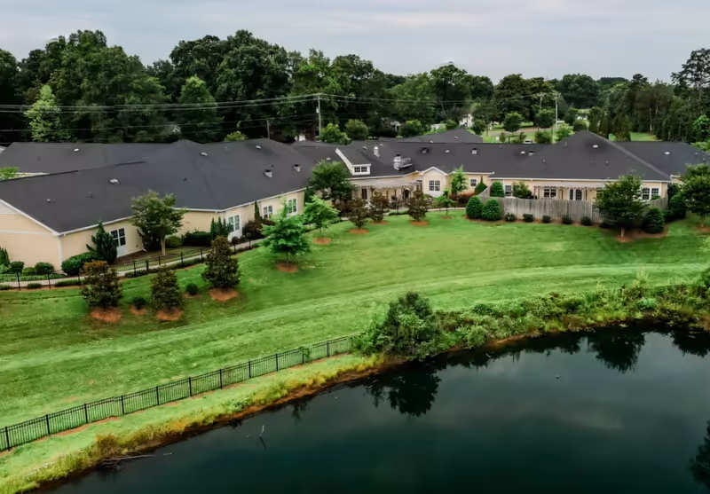 Aerial view of Cadence Senior Living at Mint Hill showing a large single-story building with a dark roof surrounded by green lawns, trees, and a pond in the foreground.
