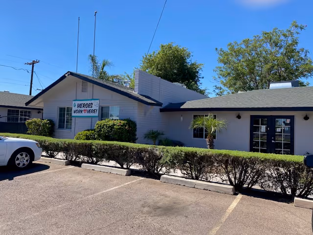 Exterior view of a single-story building with a sign that reads 'HEROES WORK HERE!' on the front wall. The building is surrounded by trimmed bushes and a parking lot with a white car parked on the left side. The sky is clear and blue.