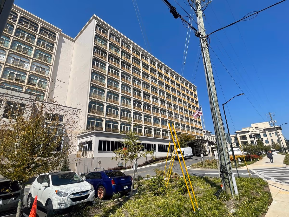 Large multi-story senior living building with rows of balconies, an American flag, parked cars and a street in front under a clear blue sky.