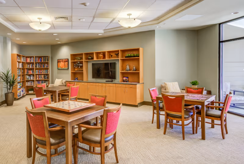 A bright and spacious common area with several wooden tables and red cushioned chairs arranged for group activities or socializing. There is a large built-in wooden entertainment center with a flat-screen TV and decorative items. A bookshelf filled with books is visible in the corner, and large windows allow natural light to fill the room. The room has a neutral color palette with beige carpeting and light-colored walls.