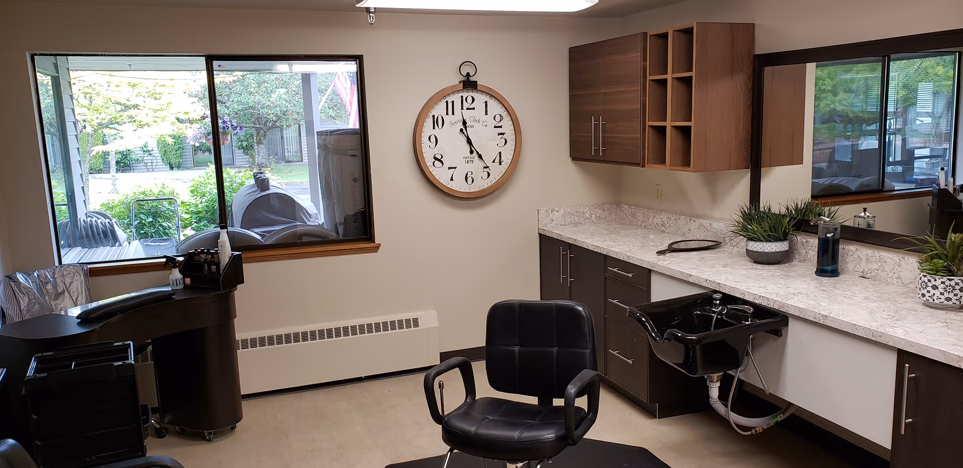 Small salon-style room with a styling chair, shampoo sink, countertop and mirror, a large wall clock, and a window looking out to a patio.