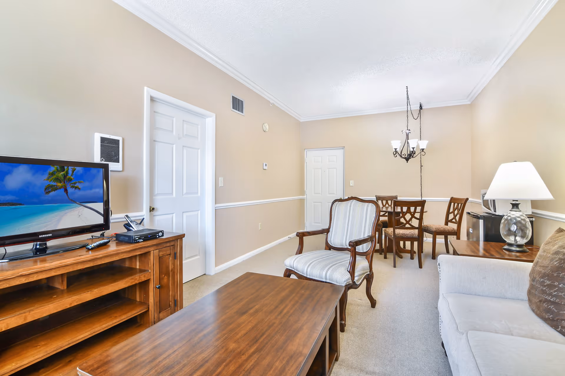 A cozy living room and dining area with beige walls and carpeted floor. The living room features a wooden coffee table, a striped upholstered armchair, a beige sofa with a brown pillow, and a wooden TV stand with a flat-screen TV displaying a tropical beach scene. The dining area has a wooden table with four chairs and a chandelier hanging above. A table lamp and a small refrigerator with a microwave on top are visible in the corner.