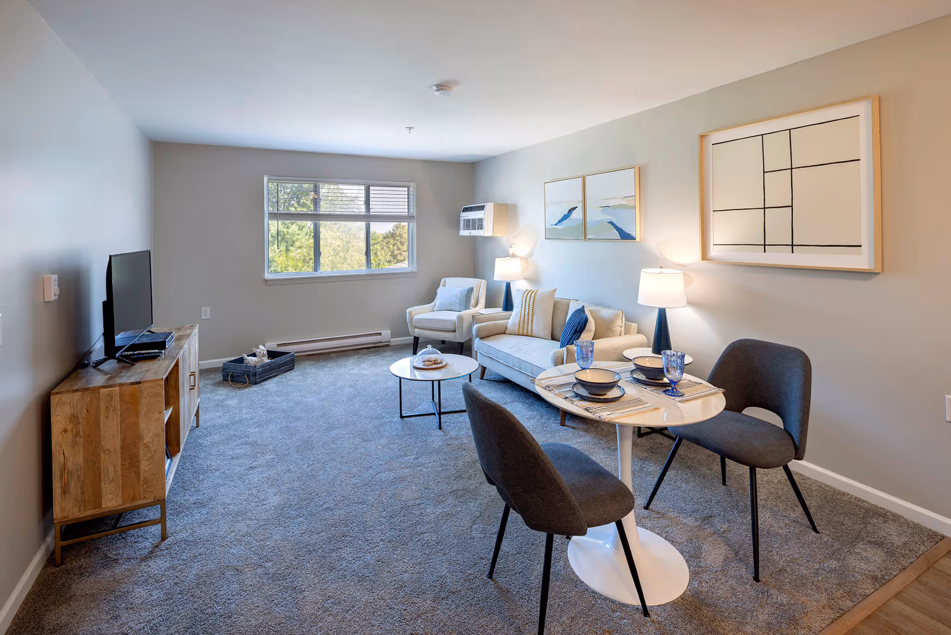 A bright living room with a large window showing greenery outside. The room features a beige sofa with cushions, a matching armchair, two table lamps, a small round coffee table with a glass dome cover, and a wooden TV stand with a flat-screen TV. There is also a small round dining table set for two with blue glasses and bowls. The walls are decorated with three framed abstract artworks.