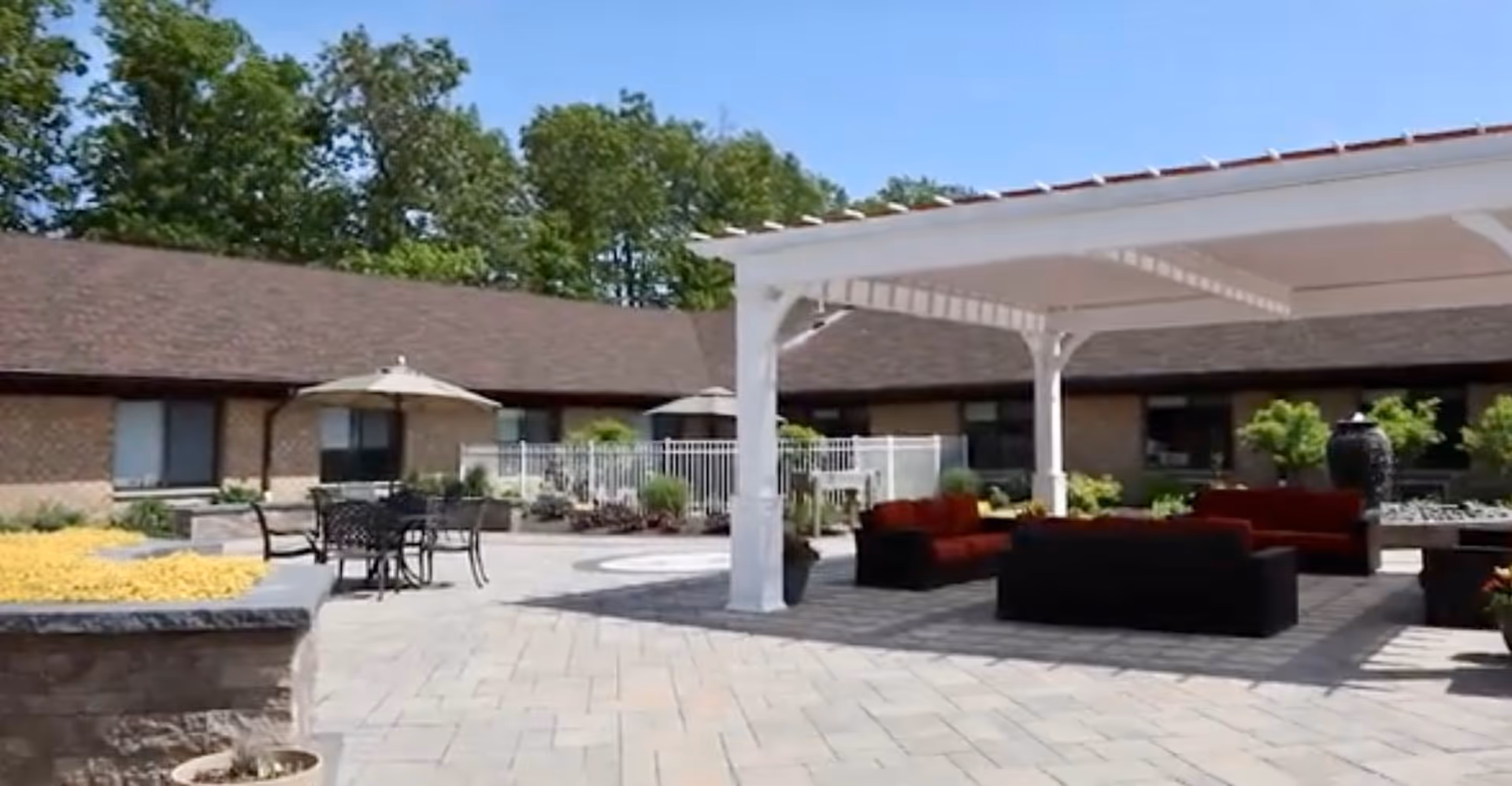 Outdoor patio area at a senior living facility with a white pergola covering red cushioned seating, black metal tables with umbrellas, surrounded by a paved stone floor and landscaping with trees and shrubs.