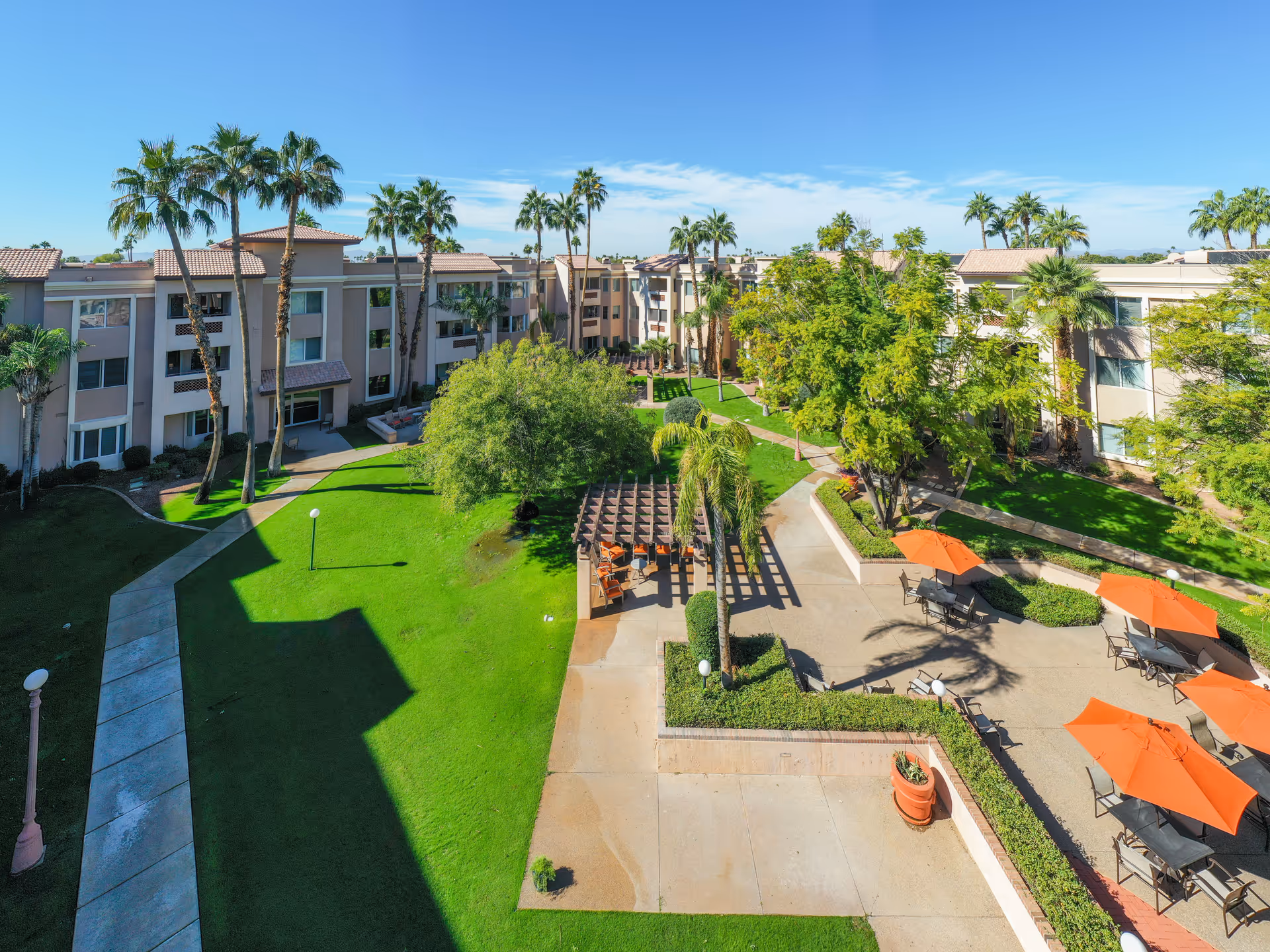 A sunny outdoor courtyard area of a senior living facility with green lawns, palm trees, and a paved patio featuring tables with orange umbrellas and chairs. The courtyard is surrounded by a three-story beige building with multiple windows and balconies.
