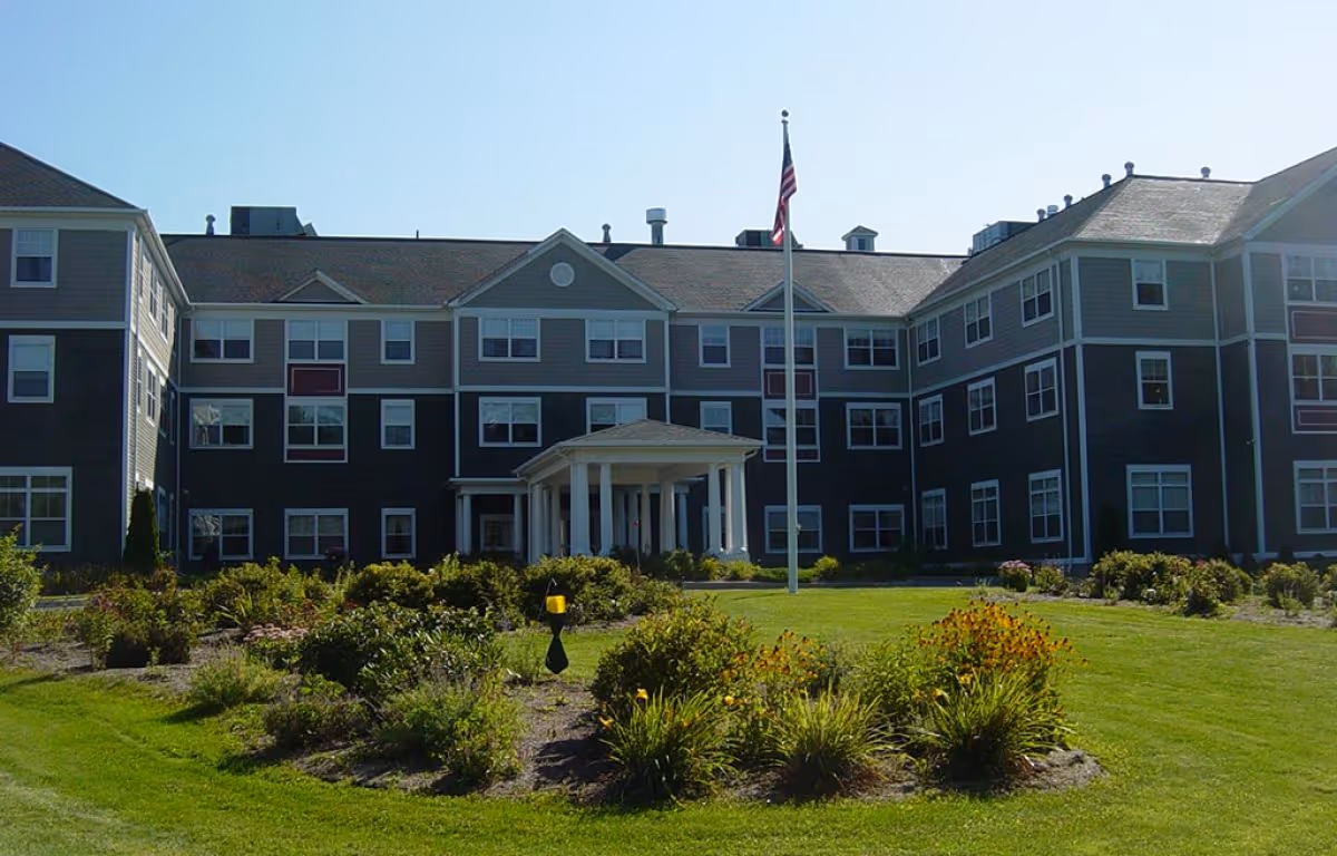 Front exterior of a three-story senior living building with a central covered entrance, flagpole, and landscaped lawn.