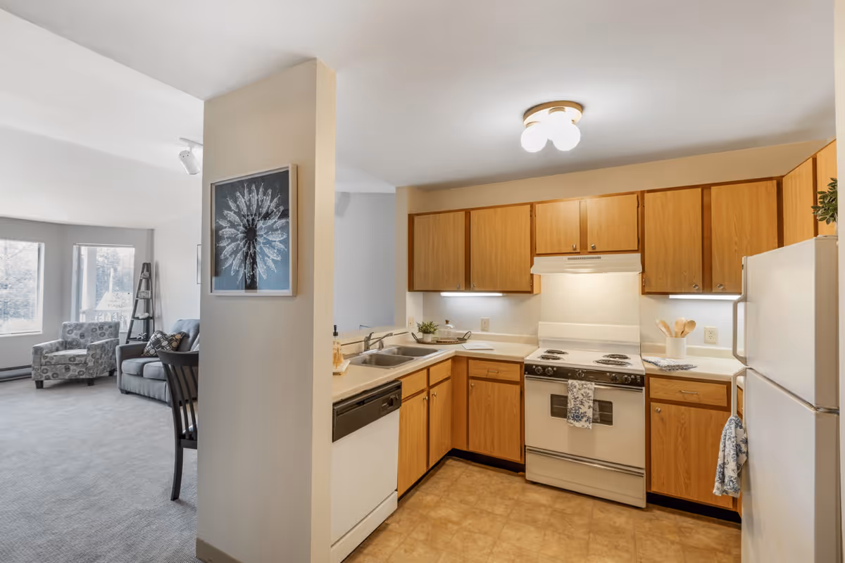 A bright kitchen area with wooden cabinets, a white stove, refrigerator, and dishwasher. The kitchen has a double sink and under-cabinet lighting. Adjacent to the kitchen is a living room with a gray sofa, patterned armchair, and large windows letting in natural light. A framed flower artwork is hung on the wall separating the kitchen and living room.