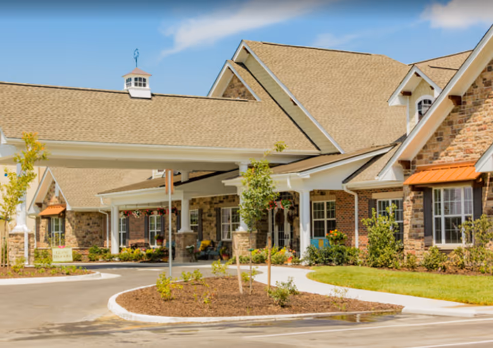 Exterior view of a senior living facility with a covered entrance, stone and brick facade, landscaped garden beds, and a clear blue sky.