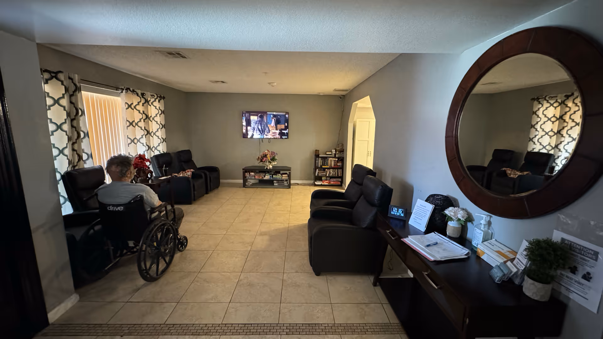 A common living area with tiled floors and light gray walls. There are several black recliner chairs arranged along the walls, a person in a wheelchair facing a wall-mounted TV, and a small table with flowers and books beneath the TV. On the right side, a dark wooden console table holds various items including a clock, papers, hand sanitizer, and a round wall mirror above it. The room has patterned curtains on the windows.