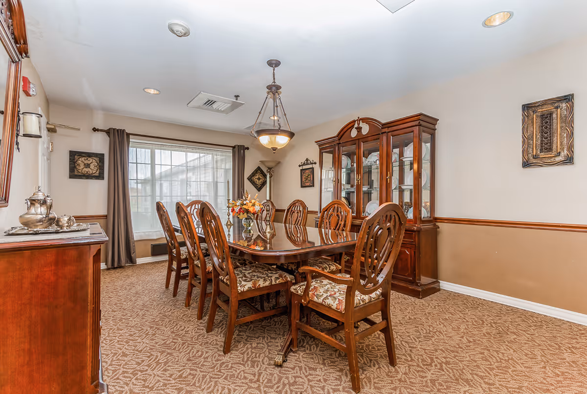 A formal dining room with a large wooden dining table surrounded by eight upholstered chairs. The room features a glass-front china cabinet filled with dishes, a sideboard with a silver tea set, beige walls with wooden trim, carpeted floor, and a large window with curtains allowing natural light to enter. A chandelier hangs above the table.