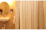 A small bathroom featuring a wall-mounted sink with a faucet and a round mirror above it. To the right, there is a shower area with a white shower curtain. The walls are tiled in a beige color, and a towel hangs on a rack next to the sink.