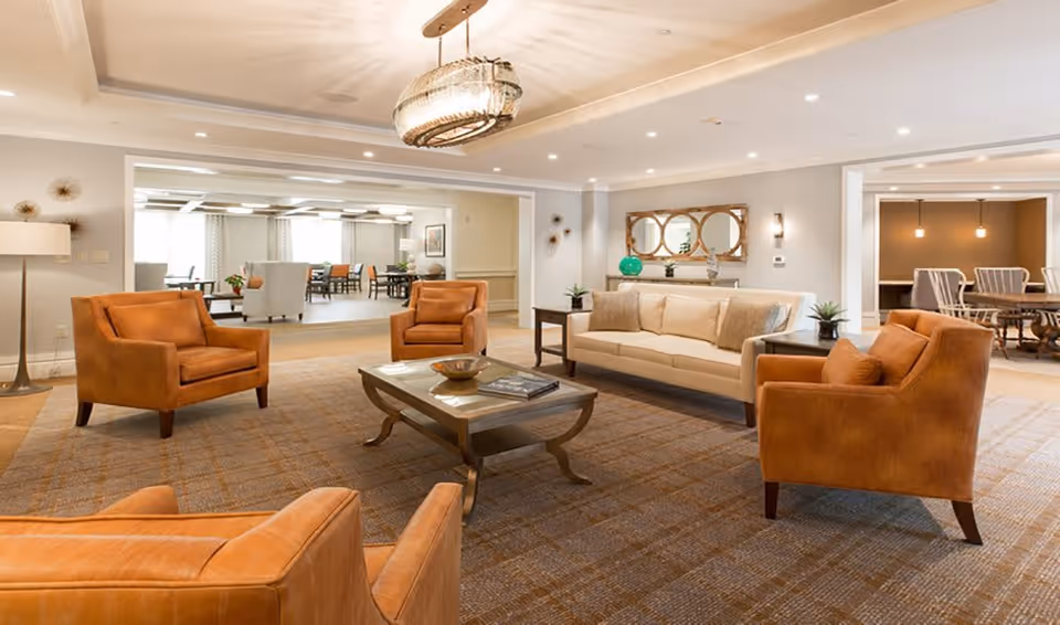 A spacious and well-lit living room area with four tan leather armchairs arranged around a wooden coffee table on a patterned carpet. A beige sofa with cushions is against the wall, beneath a decorative mirror. The room has soft lighting from ceiling fixtures and a floor lamp, with adjoining rooms visible in the background featuring dining tables and chairs.