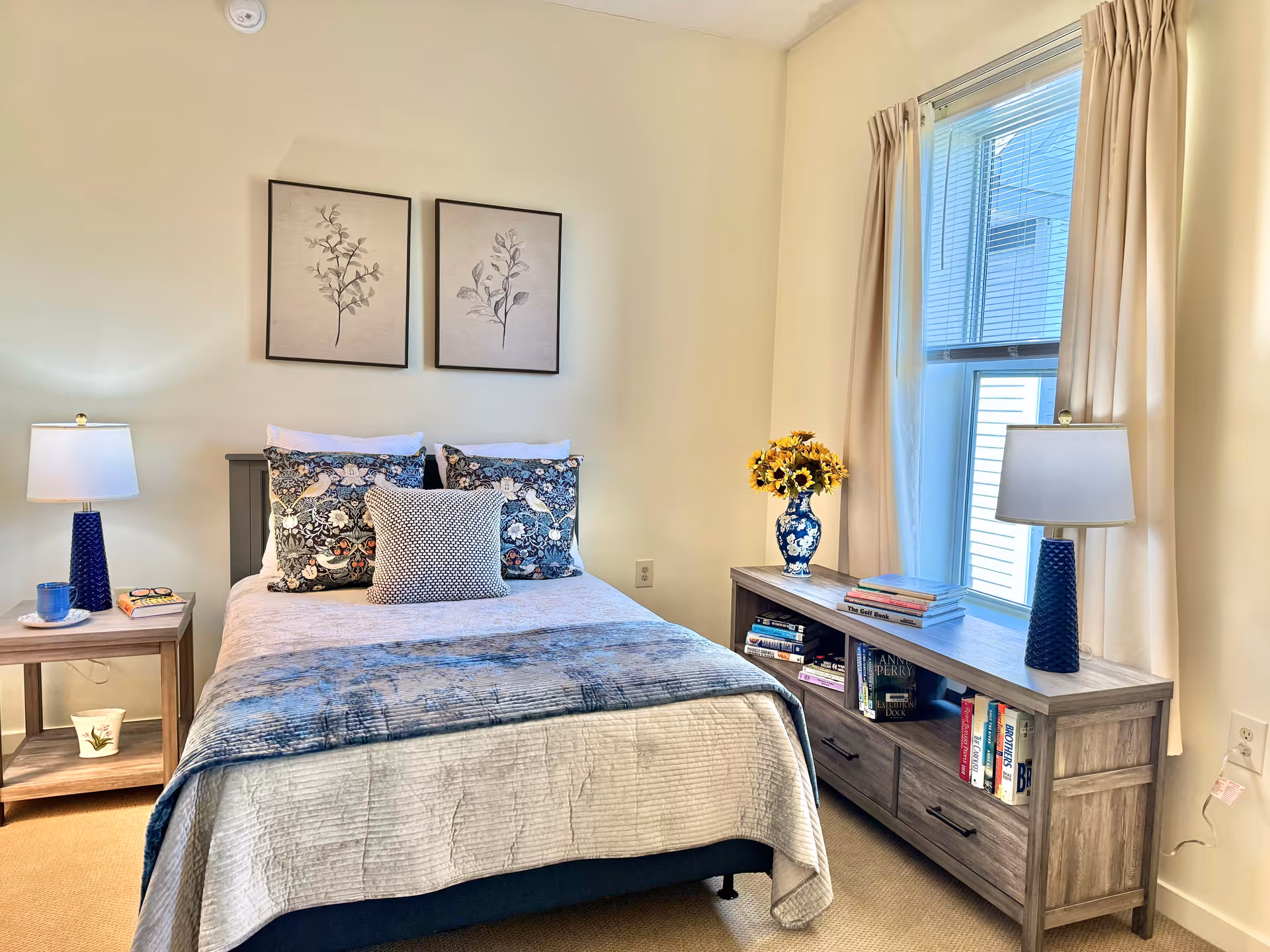 A cozy bedroom with a neatly made bed featuring patterned pillows and a blue throw blanket. On the left side of the bed is a wooden nightstand with a blue lamp, a cup, and a book. On the right side, there is a wooden dresser with shelves holding books and a blue vase with yellow flowers. Two framed botanical prints hang on the wall above the bed, and a window with beige curtains lets in natural light.