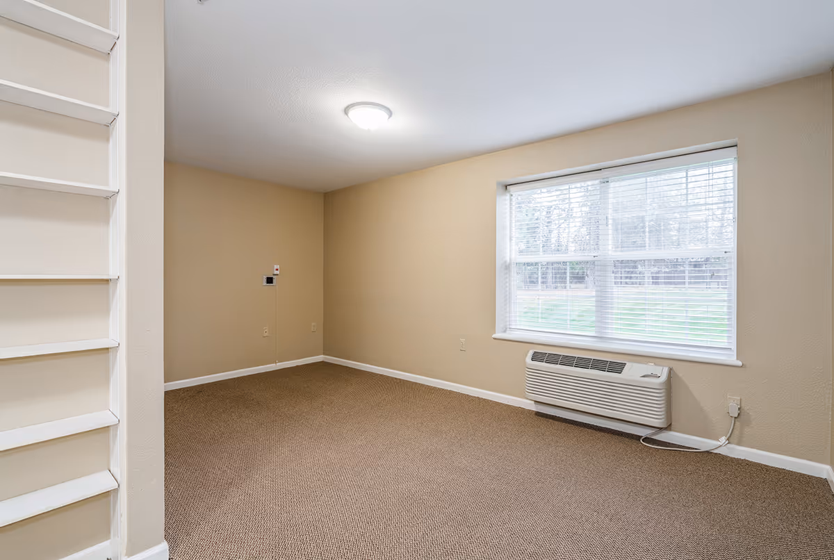 Empty room with beige walls and carpeted floor, a large window with white blinds letting in natural light, a wall-mounted air conditioning unit below the window, and a ceiling light fixture. There is a white ladder-like structure on the left side of the image.