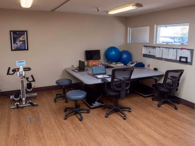 A small room with a stationary exercise bike, two blue exercise balls, a large table with three office chairs and two stools, laptops and office supplies on the table, a bulletin board with papers on the wall, and two windows showing an outdoor view.