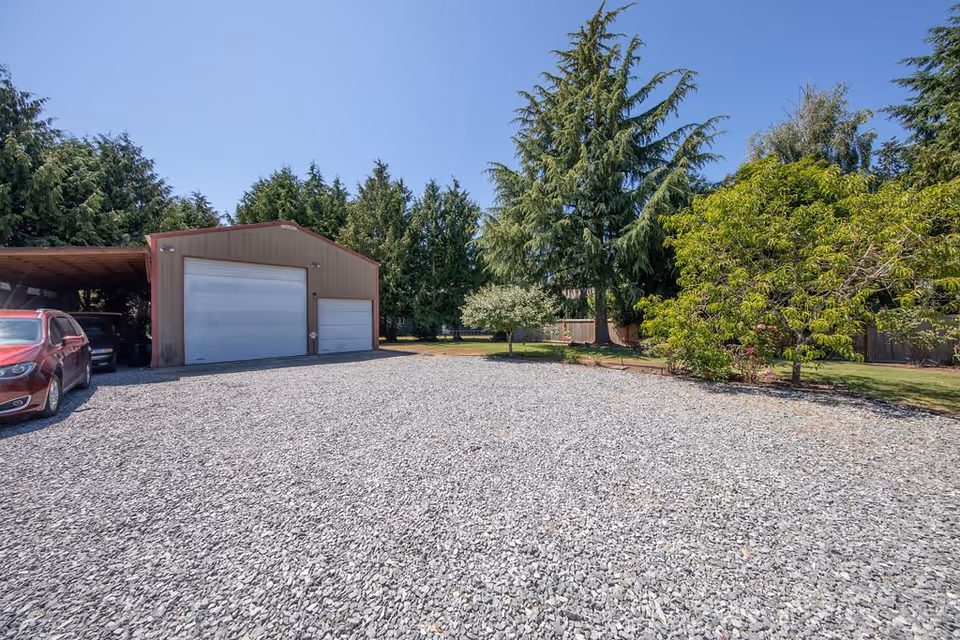 A gravel driveway leading to a large metal garage with two white doors, surrounded by tall evergreen trees and other greenery under a clear blue sky. A red car is parked on the left side near the garage.