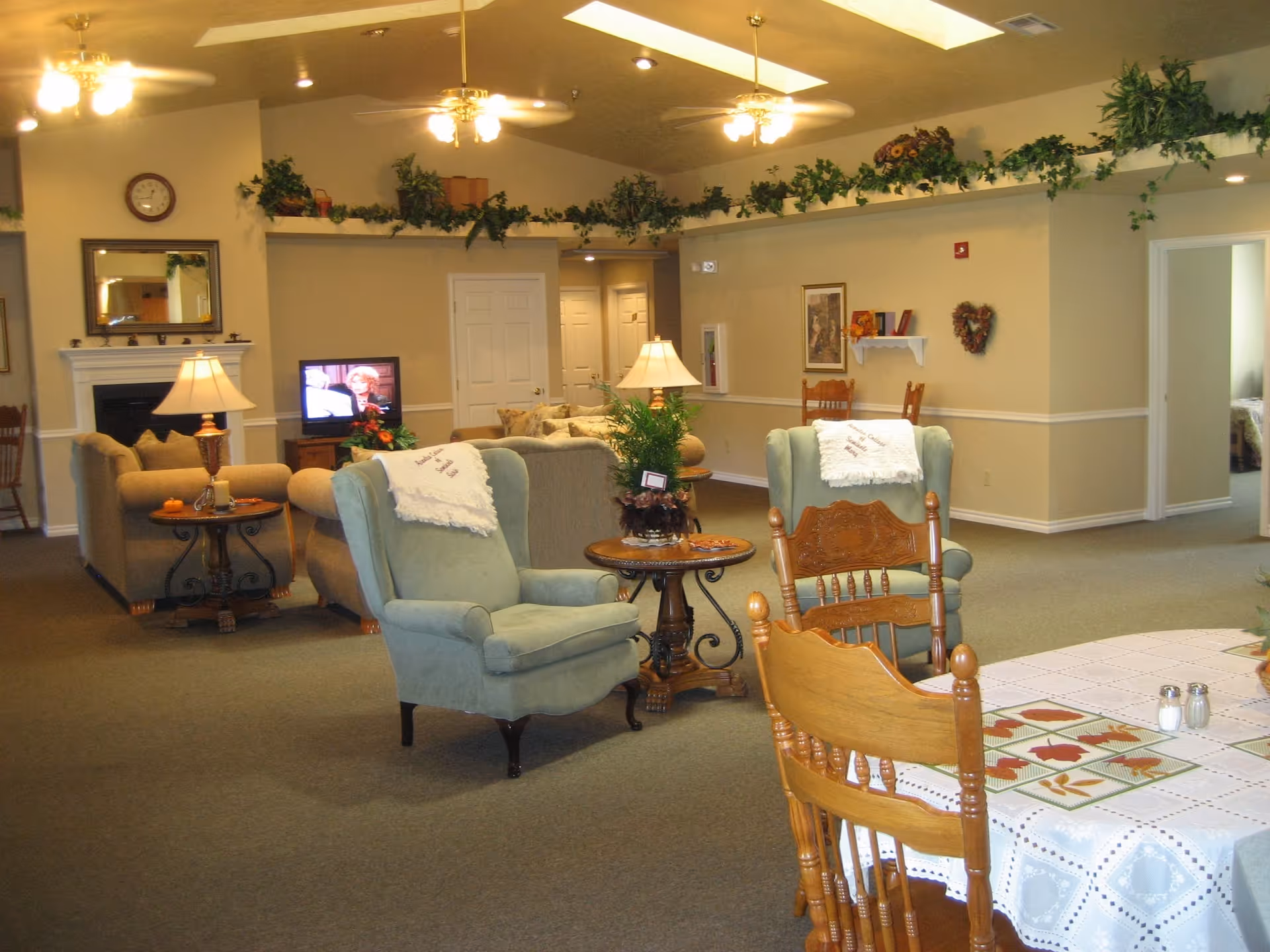 A cozy living room area in a senior living facility with comfortable armchairs, a sofa, a TV, and decorative plants on a high shelf. There is a dining table with wooden chairs in the foreground, and warm lighting from ceiling fans and lamps. The walls are beige with white trim, and there is a fireplace with a mirror above it on the left side.
