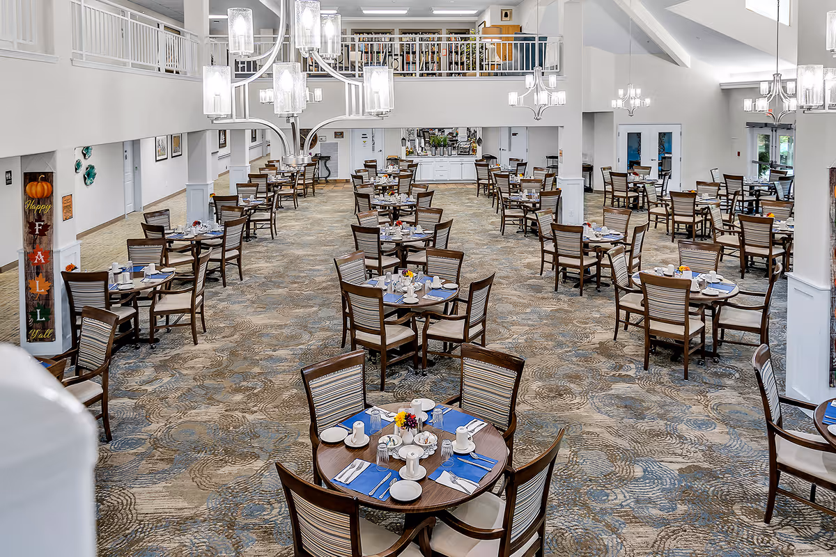 A spacious, well-lit dining room with multiple round tables set with blue placemats, white cups, plates, and silverware. Each table has four wooden chairs with cushioned seats. The room features modern chandeliers hanging from a high ceiling and a patterned carpet floor. There is a balcony area above and a decorative fall-themed sign on a pillar to the left.