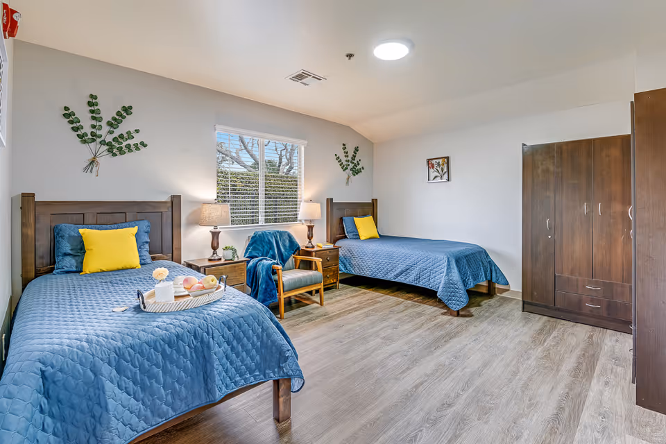 Sunny shared bedroom with two twin beds dressed in blue quilts, wooden furniture, a chair and a wardrobe.