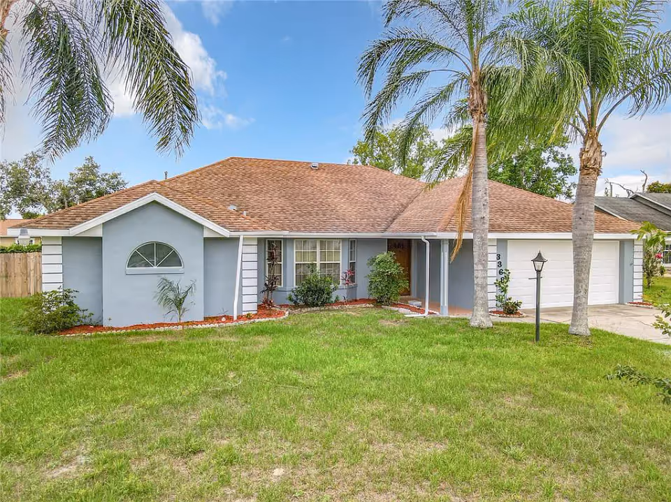 Single-story house with a brown shingle roof, light blue exterior walls, and white trim. The front yard has green grass, two tall palm trees, and a small garden bed with shrubs and plants near the house. There is a driveway leading to a two-car garage on the right side of the house. The sky is partly cloudy with blue patches.