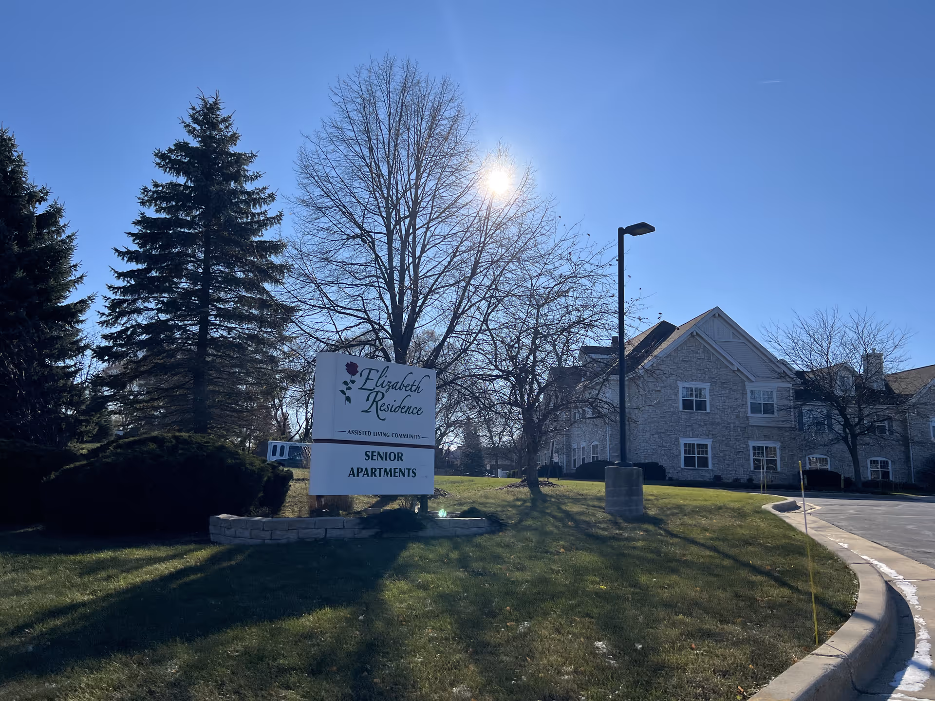 Exterior view of The Apartments At Elizabeth Residence, an assisted living community for senior apartments, showing a stone building, a sign with the facility name, trees, and a clear blue sky with the sun shining.