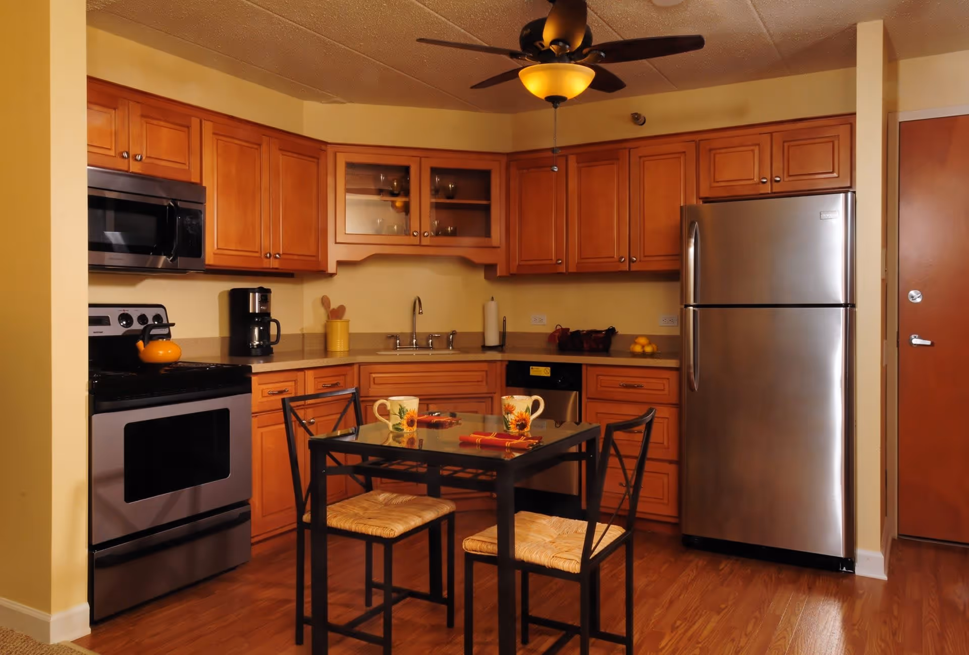 A kitchen with wooden cabinets, stainless steel appliances including a refrigerator, stove, microwave, and dishwasher. There is a small dining table with two chairs in the center, set with two mugs and napkins. The room has a ceiling fan with a light and wooden flooring.