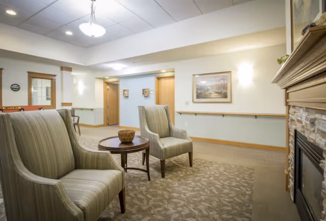 A cozy senior living facility common area with two striped armchairs and a round wooden table with a decorative bowl. The room features a carpeted floor, a fireplace with a stone surround, framed artwork on the wall, and soft lighting from ceiling fixtures and wall sconces.
