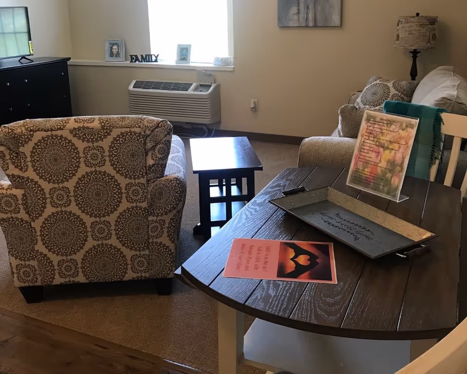 A cozy living room area with a patterned armchair, a beige sofa with decorative pillows, a wooden coffee table, and a side table with a lamp. There is a window with a heating/cooling unit below it, and a small shelf with framed photos and a decorative 'FAMILY' sign. A wooden dining table with a tray and two framed quotes is in the foreground.