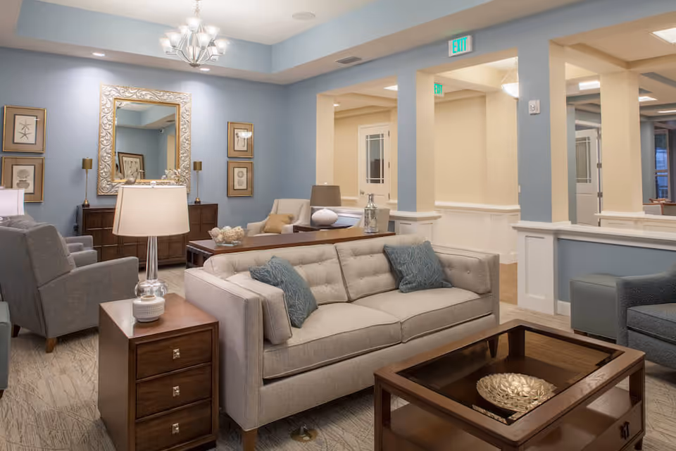 A well-lit living room with beige and gray sofas, wooden side tables, a coffee table with a decorative bowl, a large ornate mirror on a blue accent wall, framed artwork, and a chandelier overhead. The room has an open layout with beige walls and white trim.