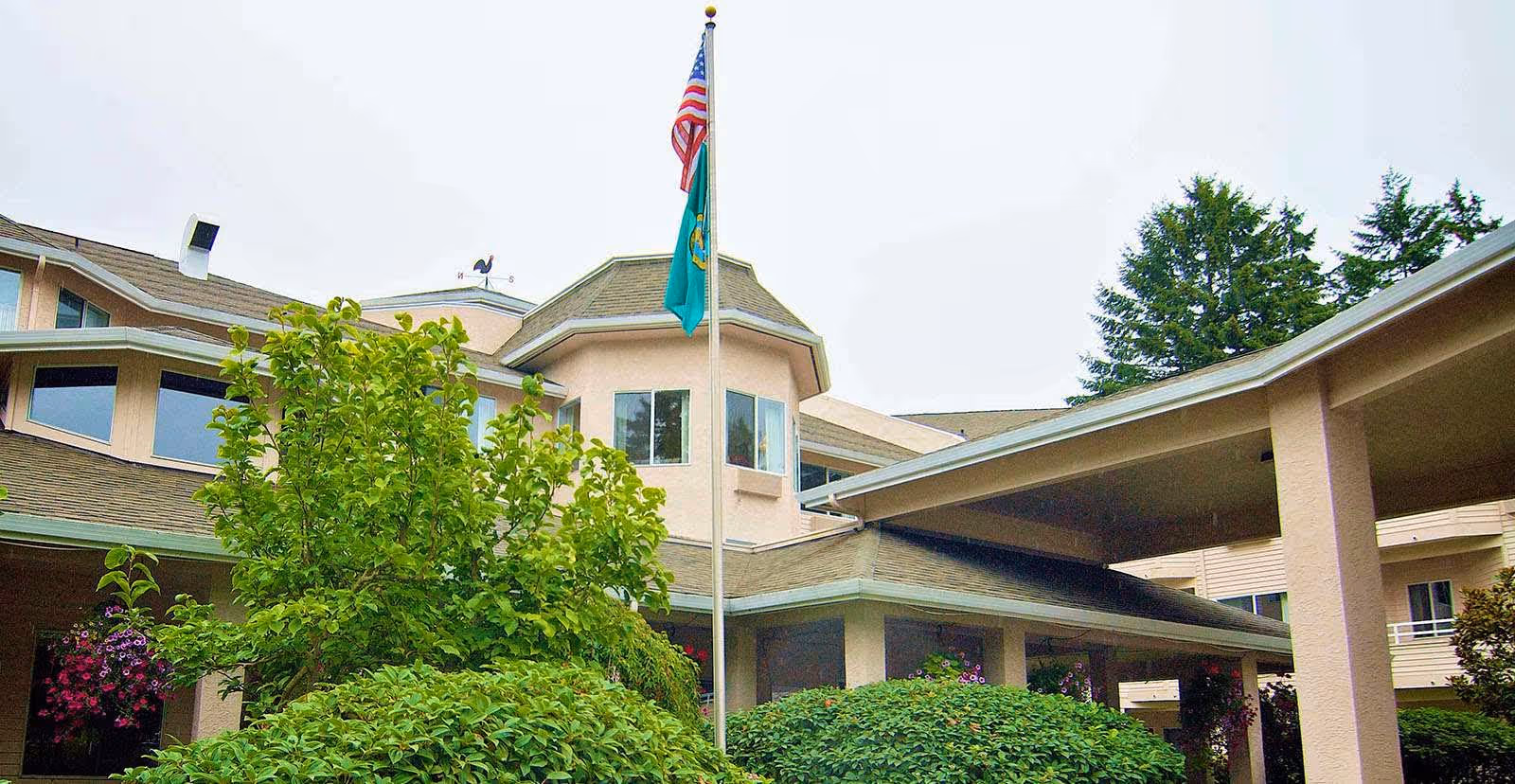 Exterior view of Solstice Senior Living at Renton showing a multi-story building with beige walls and a covered entrance. There are green bushes and trees in front, and two flags on a flagpole, one being the American flag.