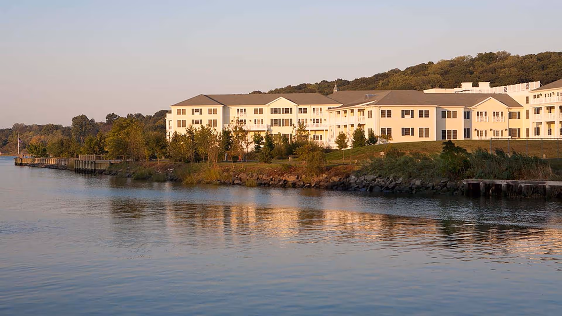 A large, multi-story senior living facility building situated along a waterfront with trees and greenery surrounding it, reflecting in the calm water during sunset.