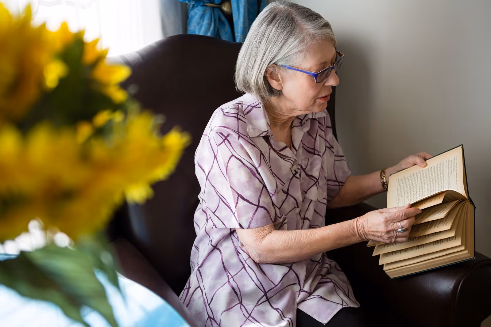 An elderly woman with gray hair and glasses is sitting in a dark armchair reading a book. She is wearing a light purple patterned blouse. In the foreground, there is a blurred bouquet of yellow flowers. The background shows a light-colored wall and a window with blue curtains.