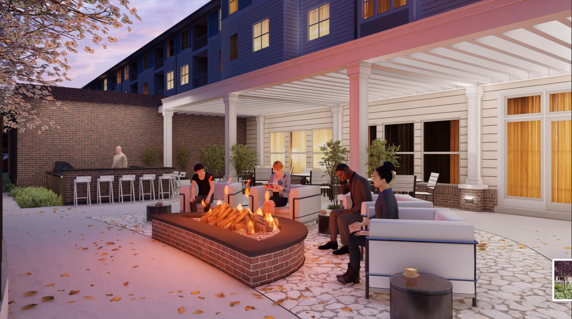 Outdoor patio area at dusk with a group of four people sitting around a rectangular brick fire pit. The patio has comfortable white cushioned seating, a bar area with stools, and is adjacent to a building with large windows and doors. There are some plants and a tree with pink blossoms visible.