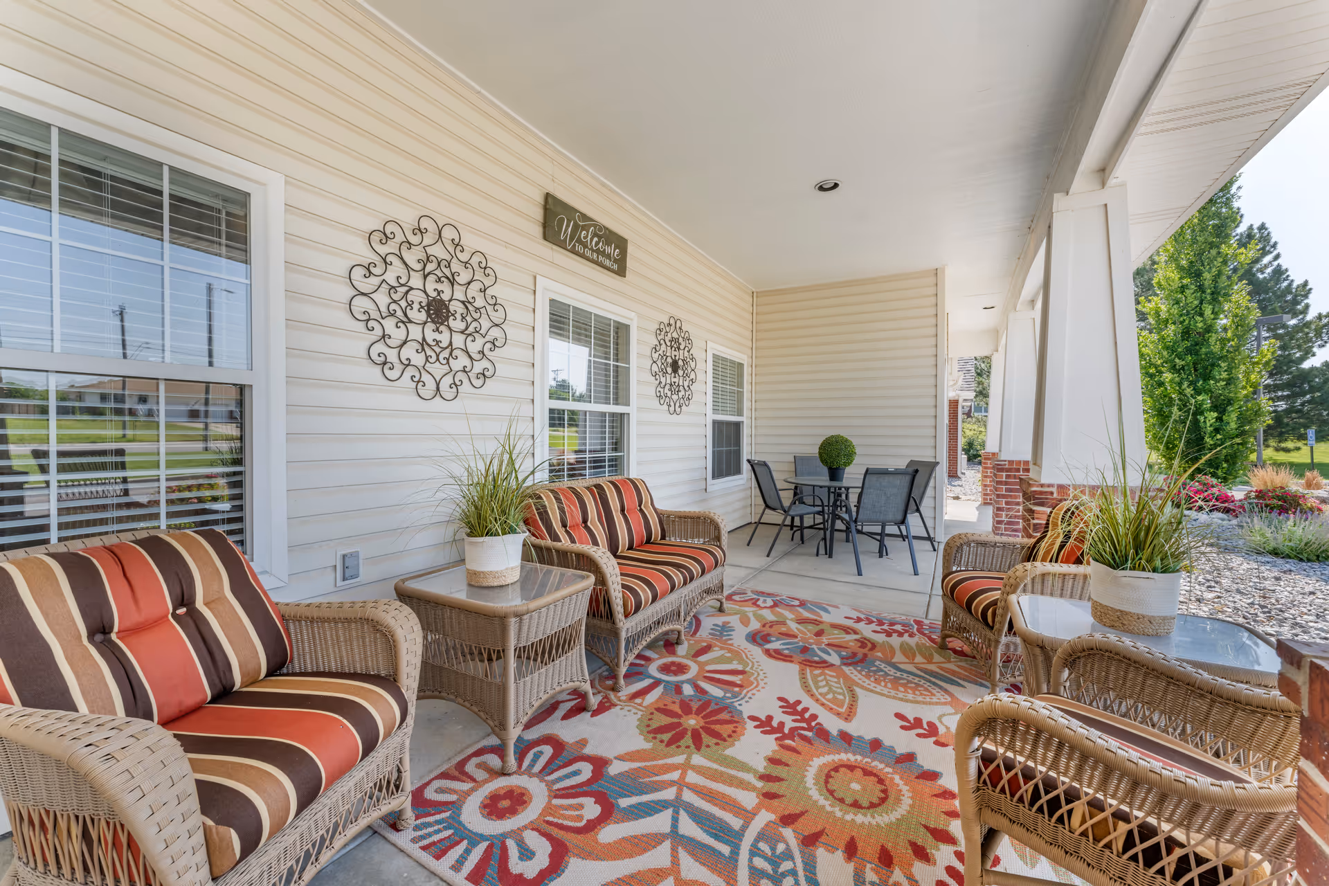 Covered outdoor porch area with wicker furniture including cushioned chairs and a loveseat with striped cushions, a colorful floral patterned rug, side tables with potted plants, a round table with four chairs, decorative wall hangings, and a sign that reads 'Welcome to our porch'.