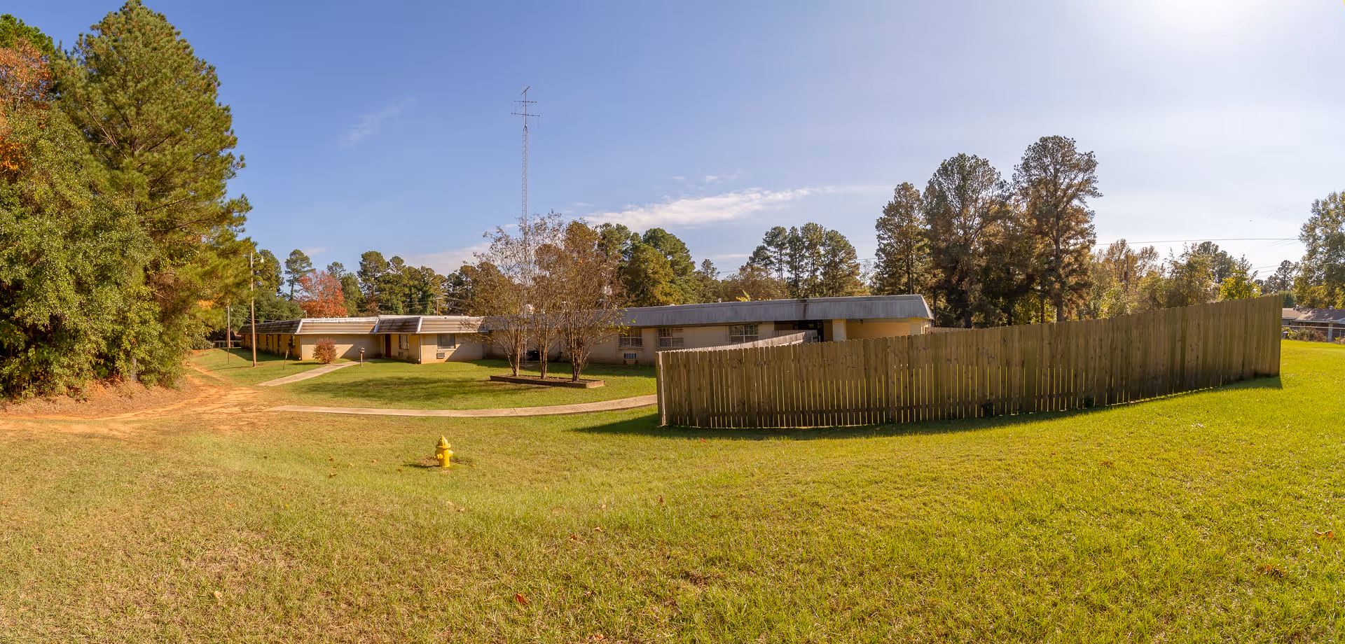 Wide view of a single-story building surrounded by grass and trees under a clear blue sky. A wooden fence encloses part of the grassy area near the building, and a yellow fire hydrant is visible on the lawn. The setting appears calm and natural.