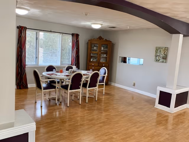 A dining area with a rectangular table surrounded by six chairs with dark cushions. The table is set with plates, cups, and utensils. There is a large window with blinds and patterned curtains on the left wall. A wooden china cabinet is placed against the back wall, and a small rectangular mirror and a decorative wall art are hung on the right wall. The floor is wooden, and the ceiling has a curved dark beam.