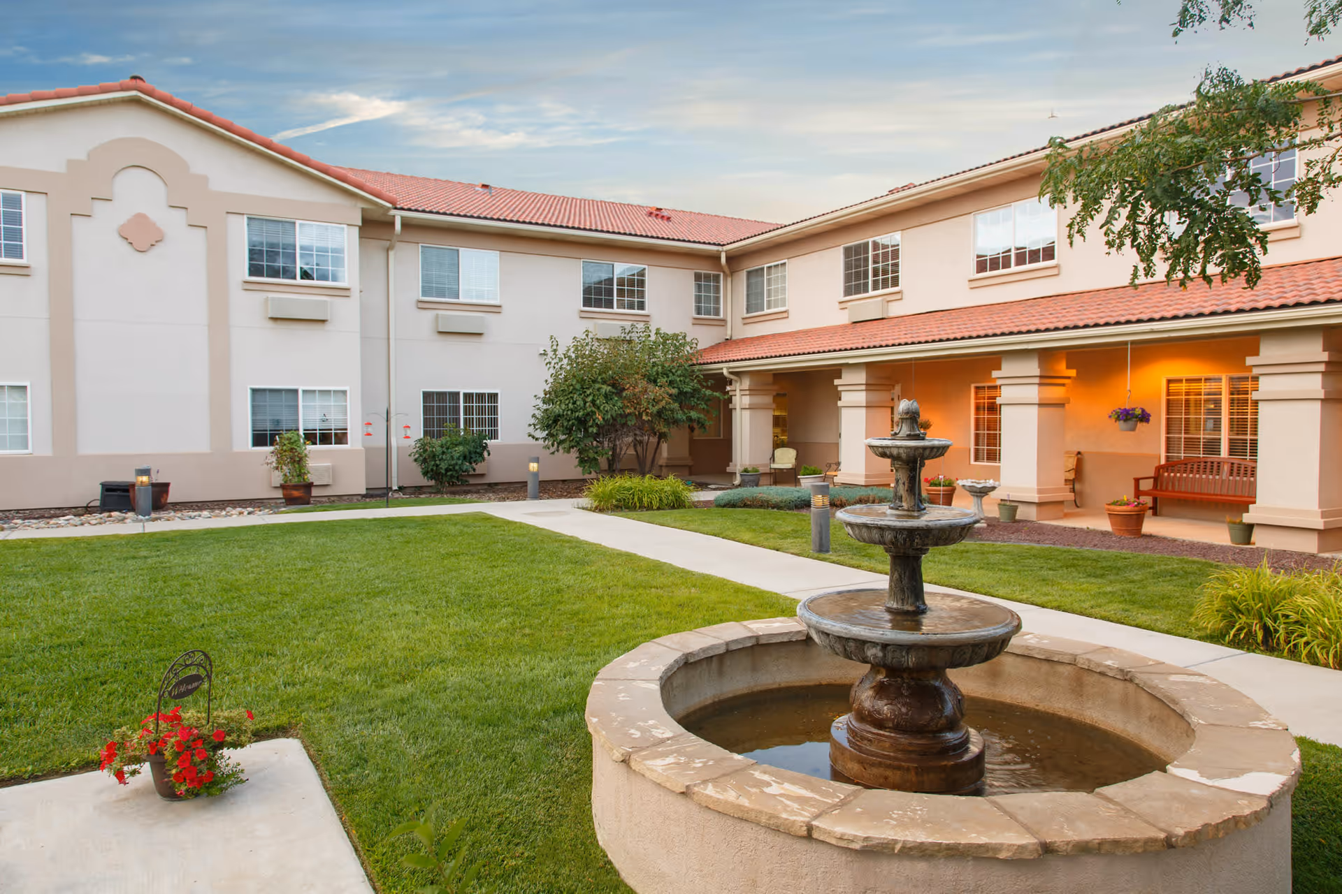 Outdoor courtyard area of The Bridge at Farmington Assisted Living featuring a stone water fountain in the foreground, green lawn, paved walkways, potted plants, and a two-story building with windows and a covered porch with seating.