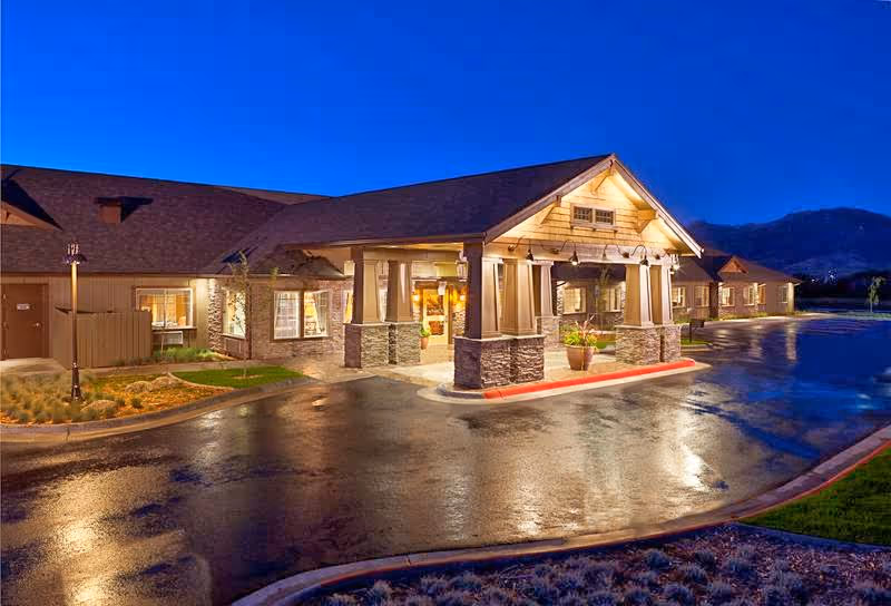 Well-lit entrance of a single-story memory care building at dusk with a wet driveway and mountains in the background.