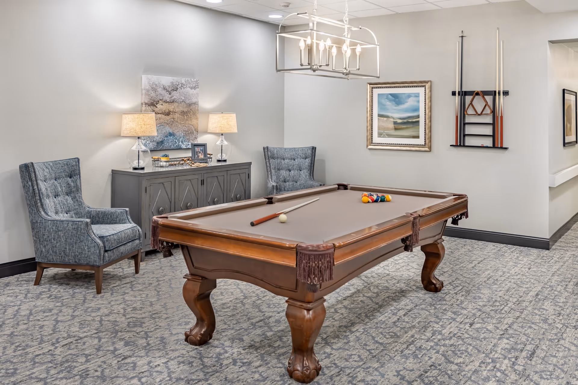 A well-lit room with a brown pool table in the center, featuring a cue stick and a rack of billiard balls. Behind the pool table, there are two blue upholstered armchairs flanking a gray sideboard with two table lamps and decorative items. On the wall above the sideboard is a piece of abstract art, and on the adjacent wall, there is a framed landscape painting and a rack holding pool cues and a triangle rack. The room has patterned carpet flooring and neutral-colored walls.