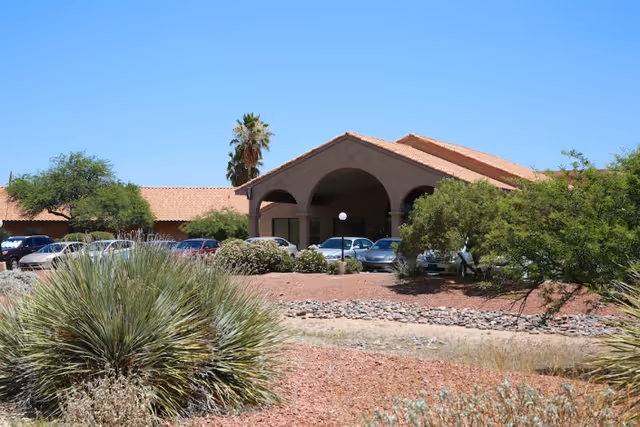 Exterior view of La Cañada Care Center building with a tiled roof, arched entrance, surrounded by desert landscaping including bushes, rocks, and a palm tree under a clear blue sky. Several cars are parked near the entrance.