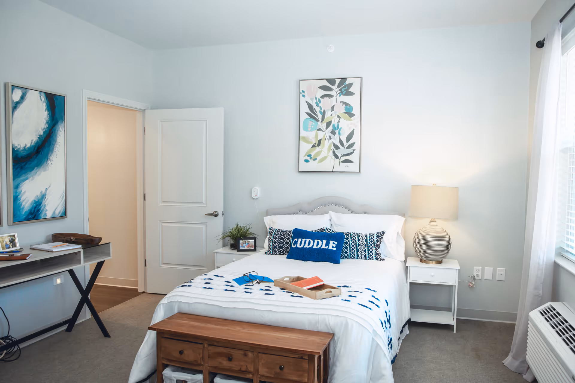 A bright and cozy bedroom with a neatly made bed featuring white and blue bedding and pillows, including one with the word 'CUDDLE'. There is a wooden bench at the foot of the bed, a white nightstand with a lamp on the right side, and a small plant and photo frame on the left nightstand. A framed botanical artwork hangs above the bed. To the left, there is a desk with some items on it and a blue abstract painting on the wall. A door is open, revealing a hallway. A window with white curtains and an air conditioning unit are on the right side of the room.