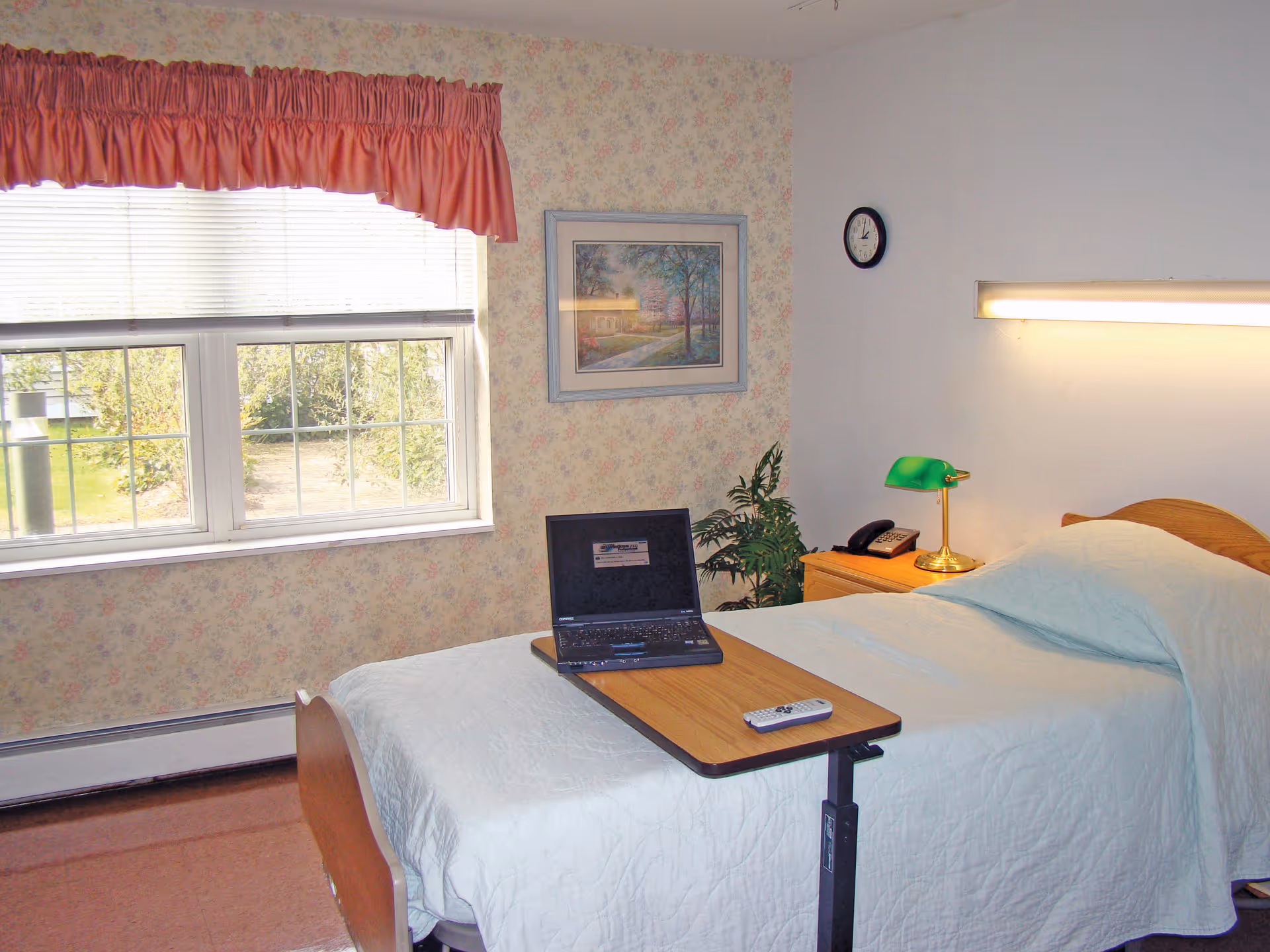 A single bed with a white bedspread in a room with floral wallpaper. A wooden overbed table holds a laptop and a remote control. Next to the bed is a wooden nightstand with a green desk lamp and a telephone. A window with white blinds and a pink valance lets in natural light. A framed picture hangs on the wall above a small plant, and a clock is mounted on the adjacent wall.
