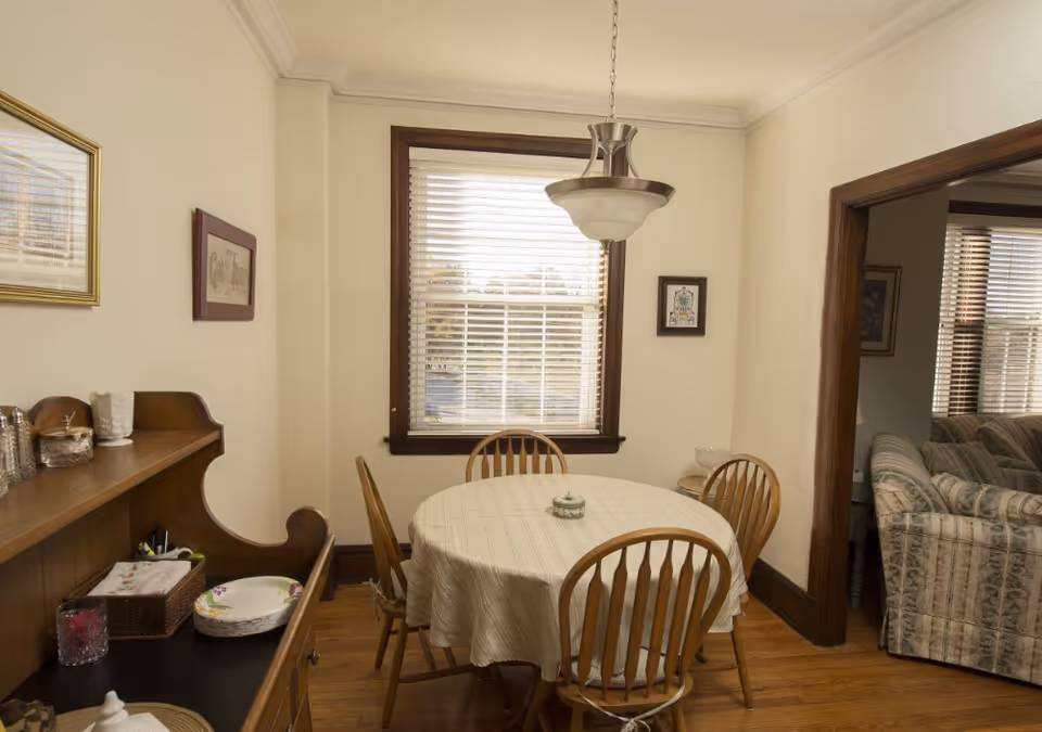 A cozy dining room with a round table covered by a beige tablecloth surrounded by four wooden chairs. A window with blinds is centered on the far wall, letting in natural light. To the left, there is a wooden sideboard with various items including plates and containers. A hanging light fixture is above the table. An open doorway on the right leads to a living room with a patterned sofa and window blinds.