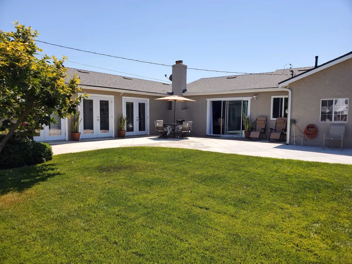 Outdoor patio area of a residential facility with a green lawn in the foreground, a tree on the left, and a beige building with multiple glass doors and windows. There is a round table with an umbrella and chairs in the center, and two reclining chairs near a sliding glass door on the right.