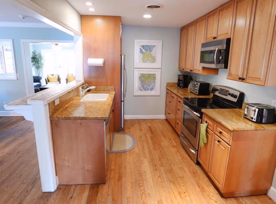 A bright kitchen with wooden cabinets, granite countertops, and hardwood floors. The kitchen features a stainless steel oven, microwave, refrigerator, toaster, and coffee maker. There is a sink with a faucet on the left countertop and two framed botanical prints on the wall. The kitchen opens into a living area with a window and seating visible in the background.