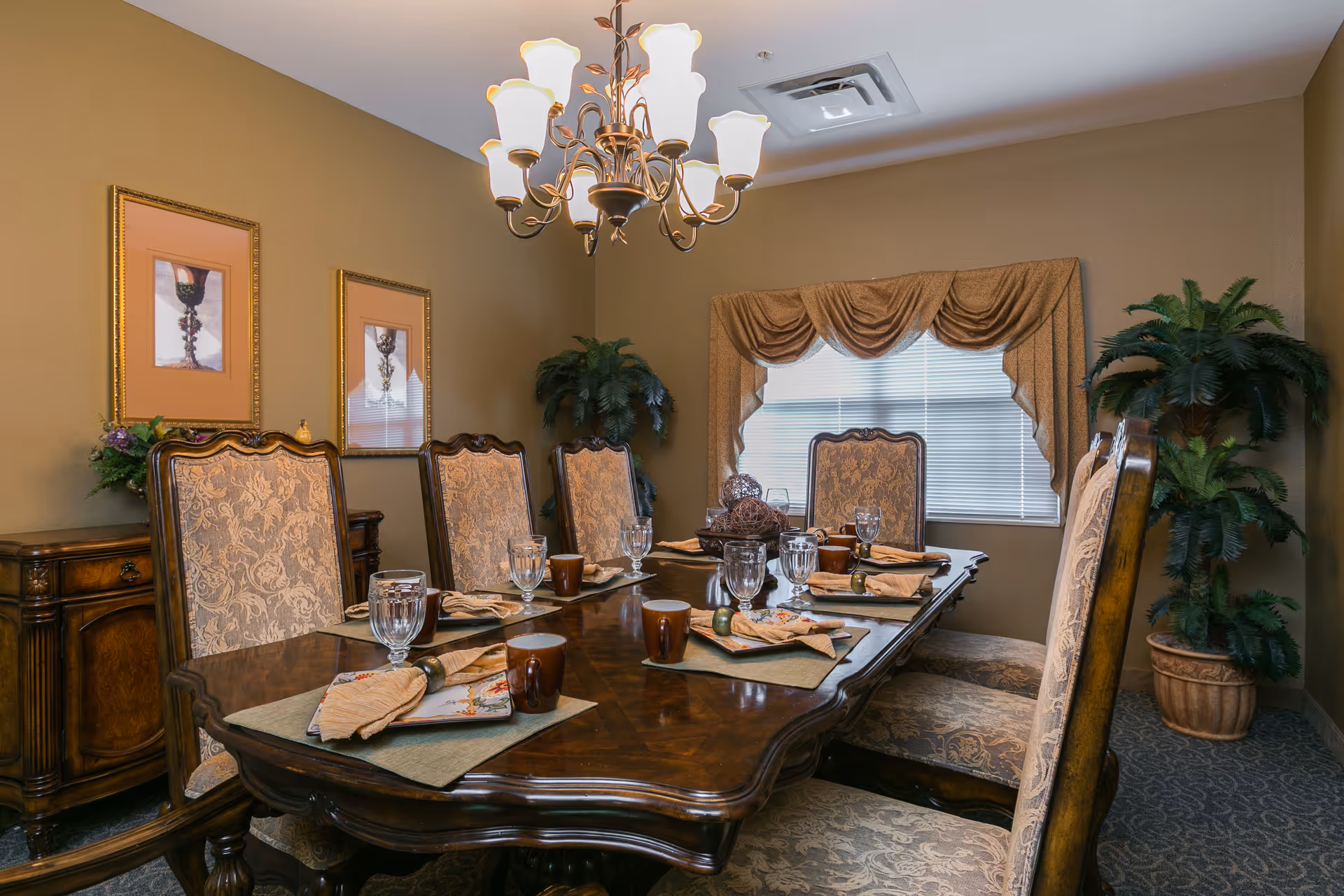 A formal dining room with a large wooden dining table set for six people. The table has placemats, napkins, glassware, and brown mugs. The chairs have ornate wooden frames and patterned upholstery. The room features beige walls, two framed pictures, a chandelier with multiple lights, a window with draped curtains, and two potted plants in the corners.